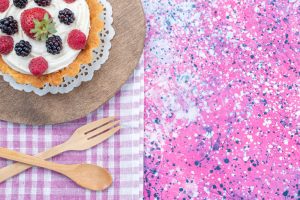 Berry-topped tart on a decorative paper doily, resting on a wooden board with a pink splatter-painted background and a plaid napkin with wooden fork and spoon nearby.