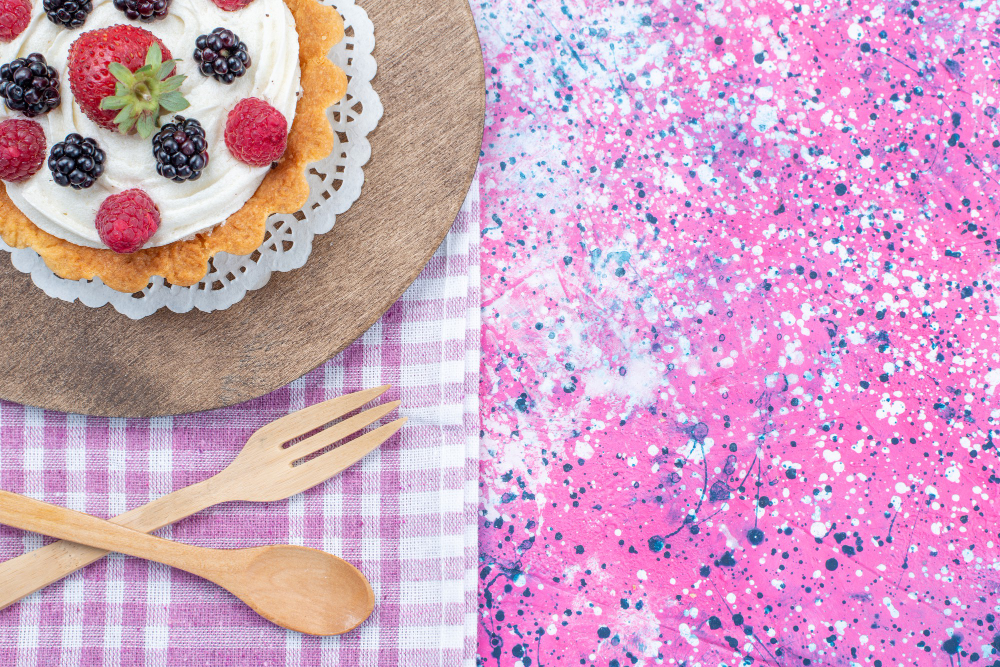 Berry-topped tart on a decorative paper doily, resting on a wooden board with a pink splatter-painted background and a plaid napkin with wooden fork and spoon nearby.