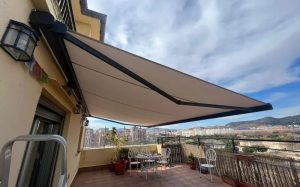 Terrace with a beige retractable awning extended, metal table and chairs, potted plants, and cityscape view in the distance.