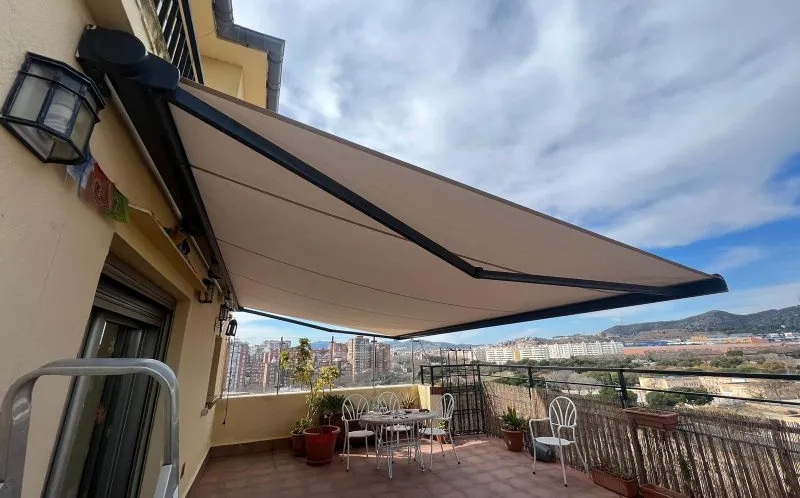 Terrace with a beige retractable awning extended, metal table and chairs, potted plants, and cityscape view in the distance.