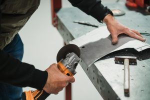Worker uses a cordless angle grinder to cut a sheet of metal on a steel workbench, hammer resting nearby on the bench edge.