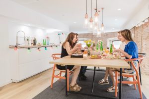 Group of friends sharing a meal at a wooden table in a bright modern kitchen with pendant lights and colorful string lights along the counter edge, plates and bottles on the table.