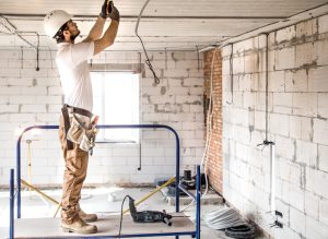 773075 - Grupo RF - Decorar.org Construction worker in a white hard hat and tool belt stands on a scaffold, wiring the ceiling in an unfinished room.