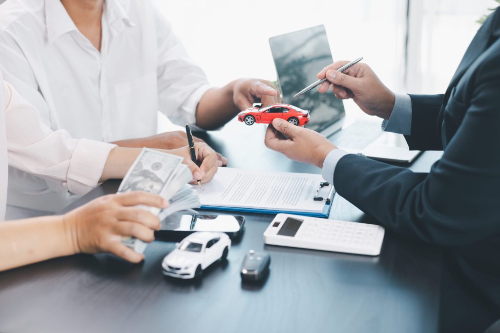 Businesspeople discuss auto financing, presenting a red toy car over papers and a calculator on the desk.