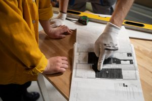 Worker in a yellow jacket points at a blueprint spread on a wooden table while another hand steadies the sheet; gloves worn for safety.
