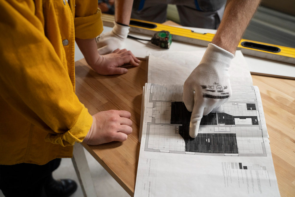 Worker in a yellow jacket points at a blueprint spread on a wooden table while another hand steadies the sheet; gloves worn for safety.
