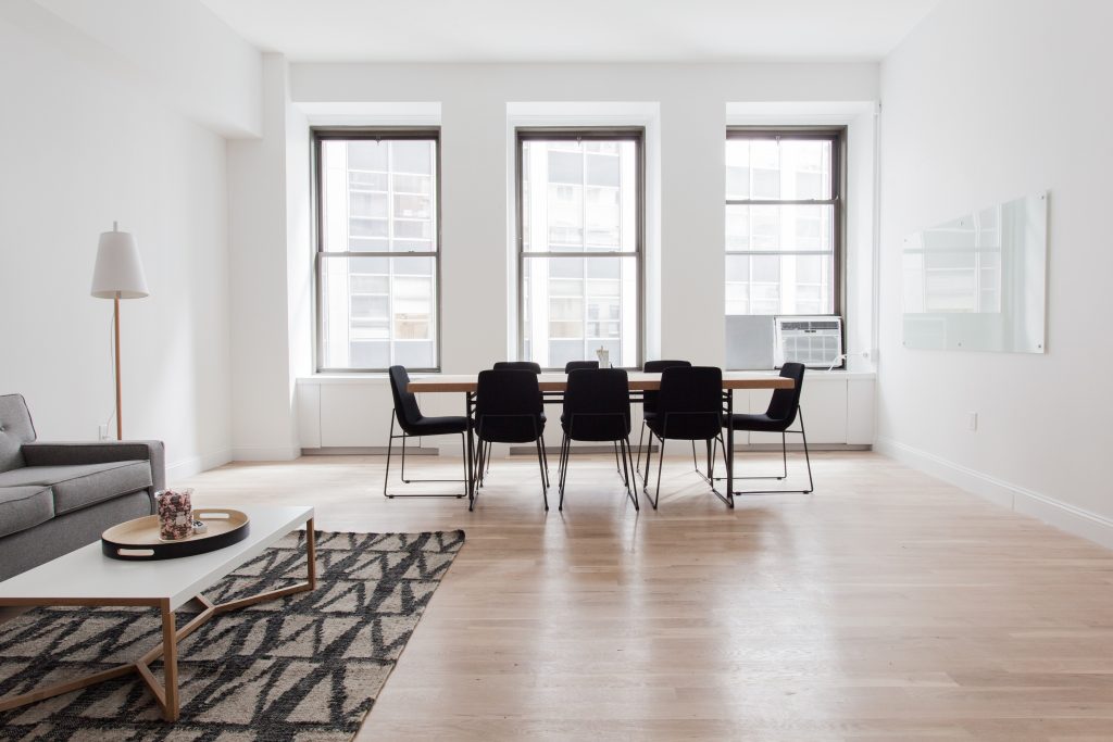 Open living/dining area with a long wooden table and eight black chairs facing three large windows; minimal furniture and light wood floor.