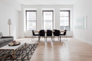 Open living/dining area with a long wooden table and eight black chairs facing three large windows; minimal furniture and light wood floor.