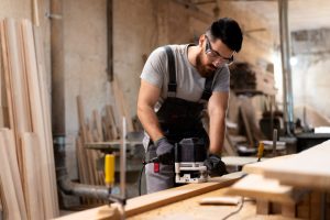 Bearded man in safety goggles and overalls guides a router along a wooden plank in a woodworking shop.