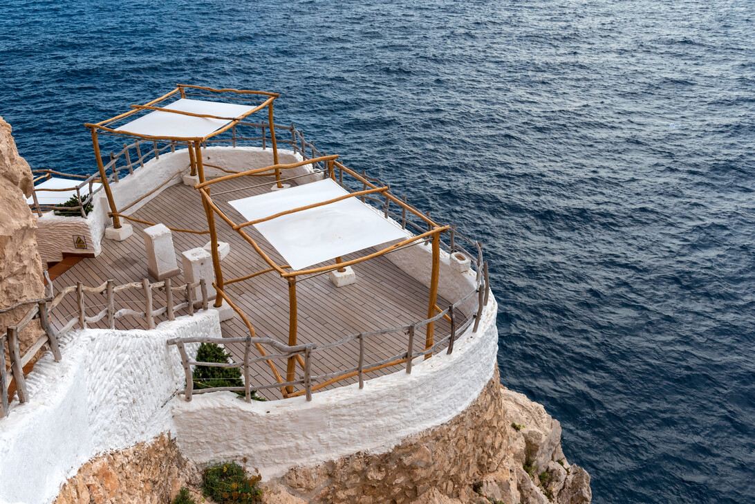 Whitewashed terrace with wooden railings on a cliff overlooking a blue sea, shaded seating canopies.