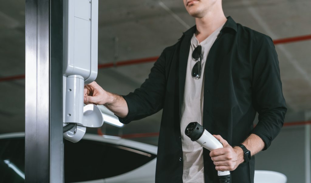 Man in a black shirt holds a white electric vehicle charging plug and approaches a charging station to connect it to a car in a garage/parking area.