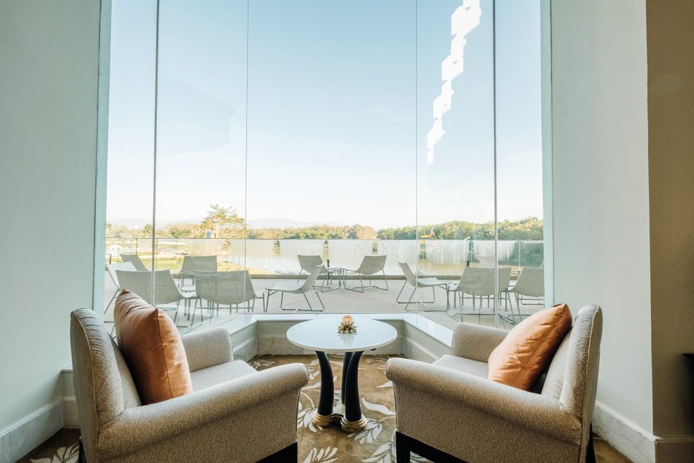 Two beige armchairs with orange throw pillows face a small round table inside a bright lounge, overlooking a lakeside patio through floor‑to‑ceiling glass walls.
