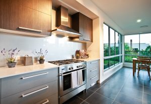 Modern kitchen with a stainless steel gas range and hood, grey cabinets, white countertops, and a bright dining nook by large windows overlooking greenery.