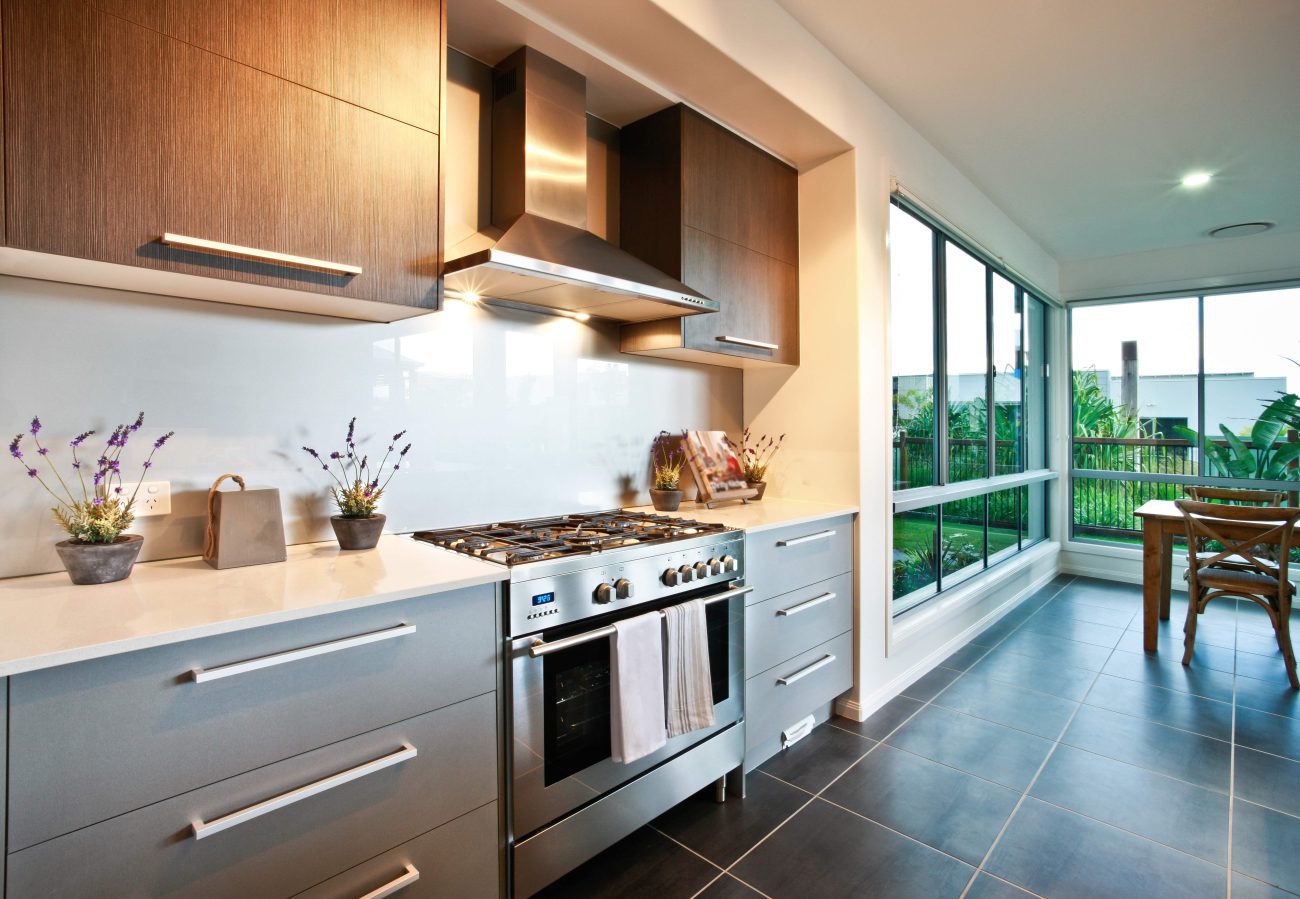 Modern kitchen with a stainless steel gas range and hood, grey cabinets, white countertops, and a bright dining nook by large windows overlooking greenery.