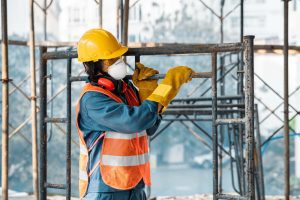 Construction worker in a yellow hard hat, orange reflective vest, gloves, and mask inspecting rusted scaffolding on a construction site.