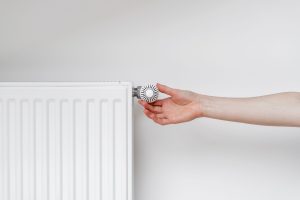 Person adjusting a radiator valve with a round knob to control heat on a white radiator wall.