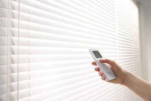 Person's hand holding a white remote control near closed horizontal window blinds, as if adjusting them
